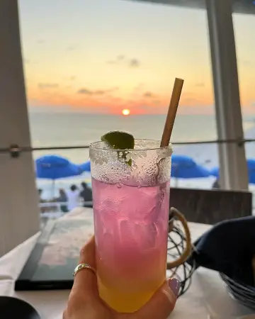 Cocktail in a woman's hand which is held out in front of the sunset out the window at Spinner's