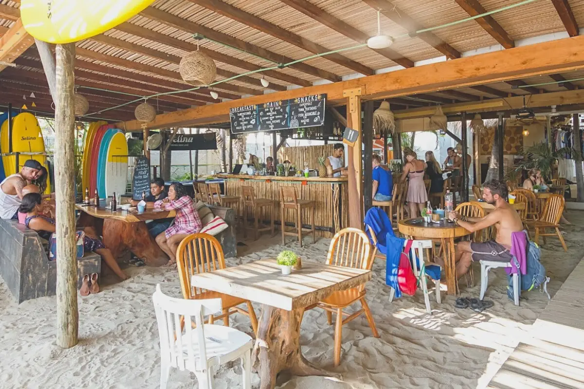 Bar and restaurant area with hostel guests hanging out and mingling with a few people at the bar, under a wooden roof and with a sand floor