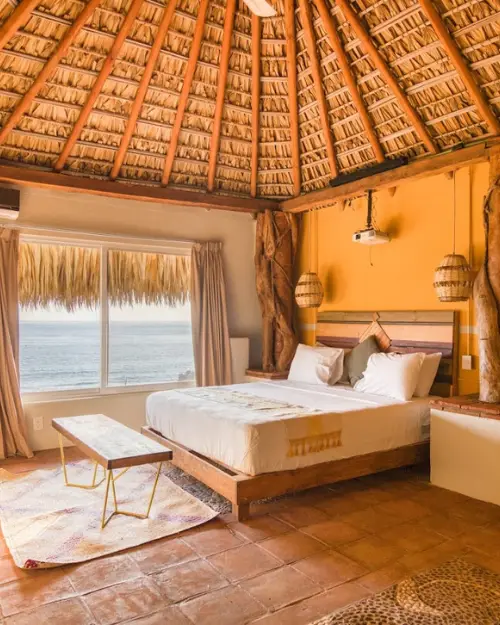 A queen sized bed in a large room with a palapa style roof next to a window overlooking the ocean