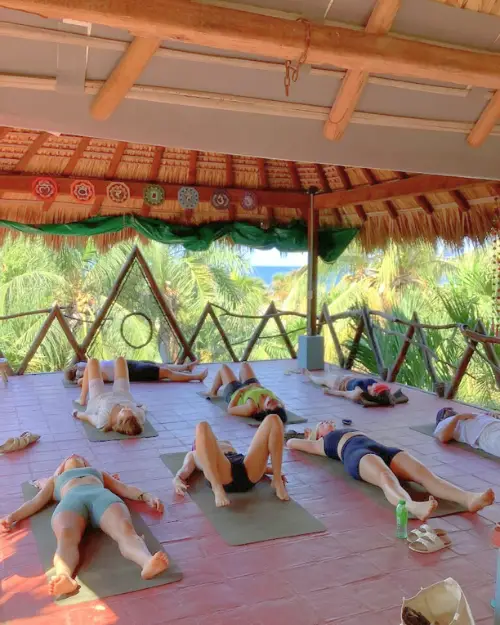 A group of people laying on their backs during a yoga class in an open air space up in the trees overlooking the ocean and palm trees