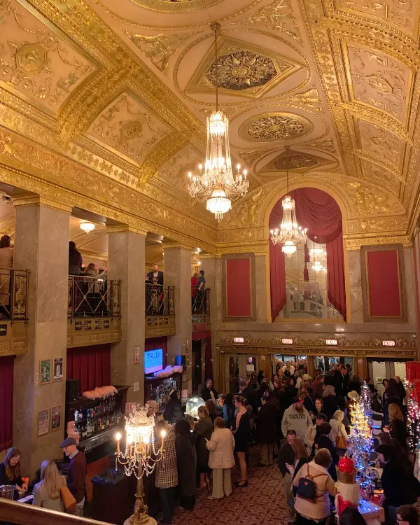 The stunning lobby of the Warner Theatre in DC, with giant chandeliers and decorated ceilings above while people get drinks and merchandise below