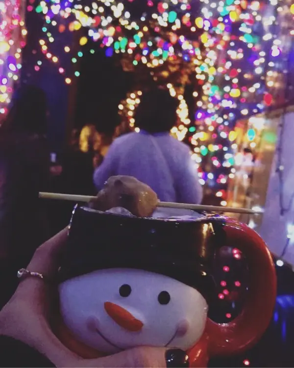 A snowman mug with a bite of cookie dough on top with holiday lights shining in the background