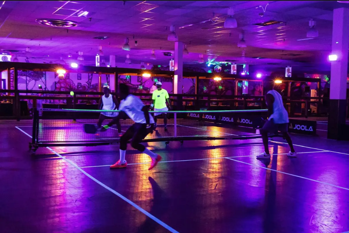 People playing pickleball with glowing shirts and headbands in a room that is glowing purple/pink