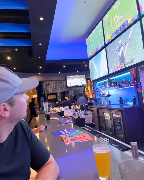 A man looking up at big screen TVs with a beer at the Dave and Busters bar