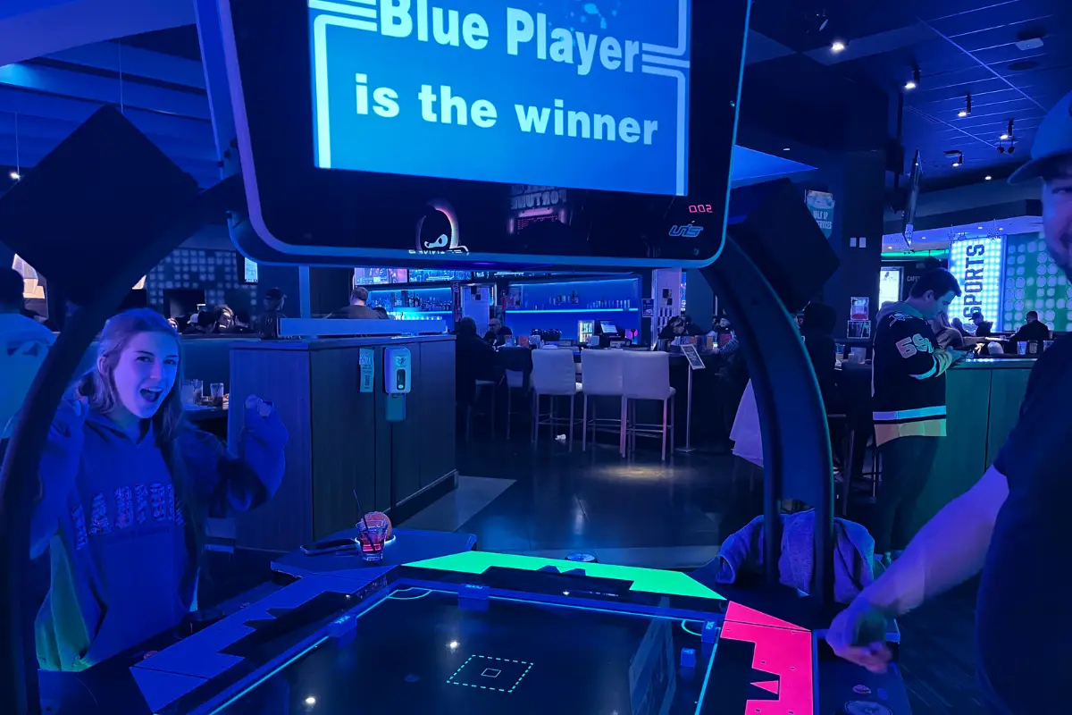 Girl excited to have won a game in an arcade with "Blue Player is the Winner!" lit up on the game screen