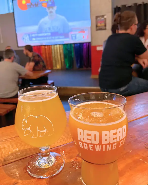 Two beers in glasses at Red Bear Brewing, one of which has a bear logo on it with sports playing on a TV in the back