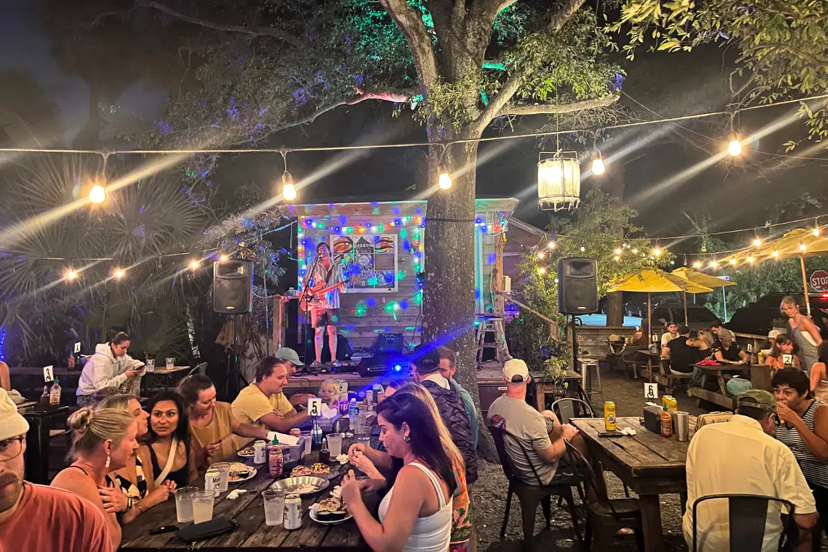 A band performing on stage under string lights and Folly Beach foliage at night at Chico Feo with people eating and enjoying during the show 