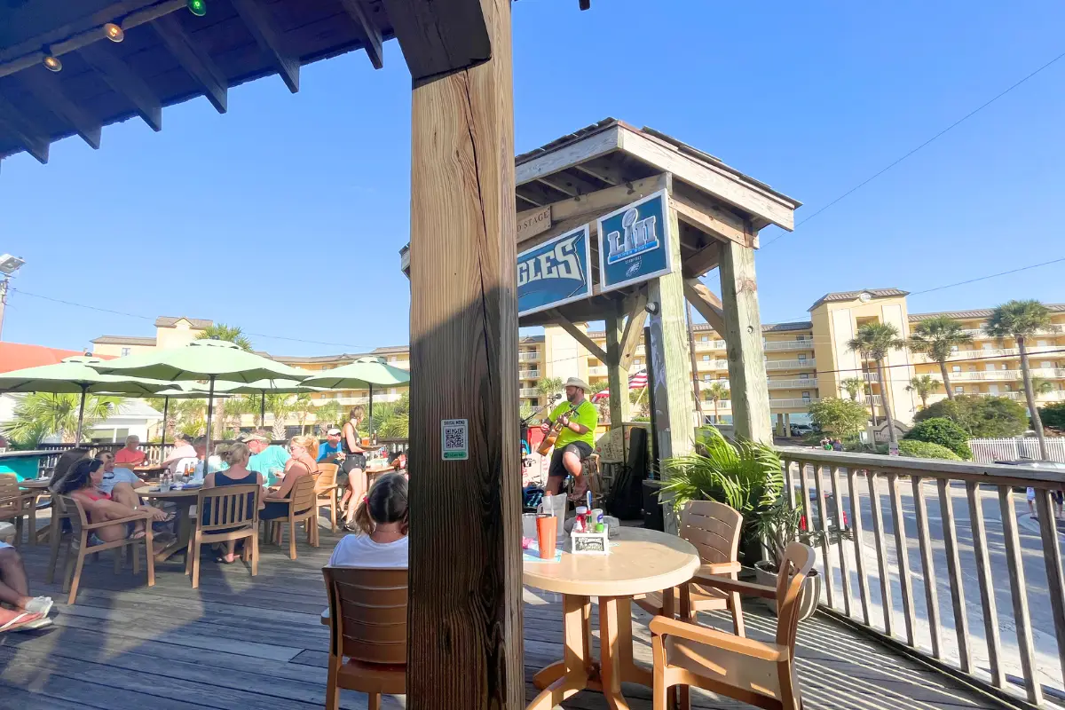 A man playing acoustic-style music on the stage at the deck at Loggerheads under a sunny sky with people happily watching, to show one of the best places to catch live music Folly Beach SC on a nice day