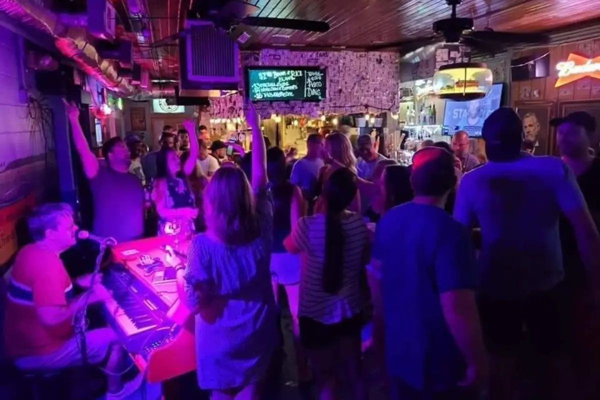 A musician playing music on a piano in a crowded dive bar with people crowded around singing and dancing under neon lights