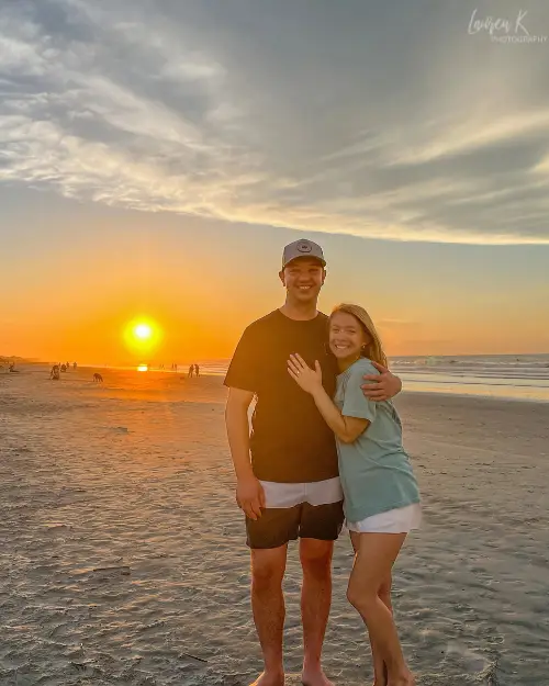 A couple smiling at the camera in front of a sunset on the beach